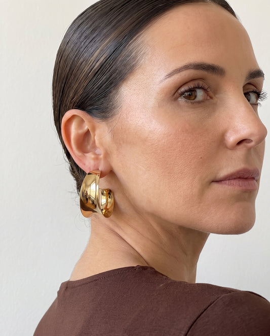 Pendientes de aro ancho dorado y curvado, puestos con camisa marrón y el pelo recogido