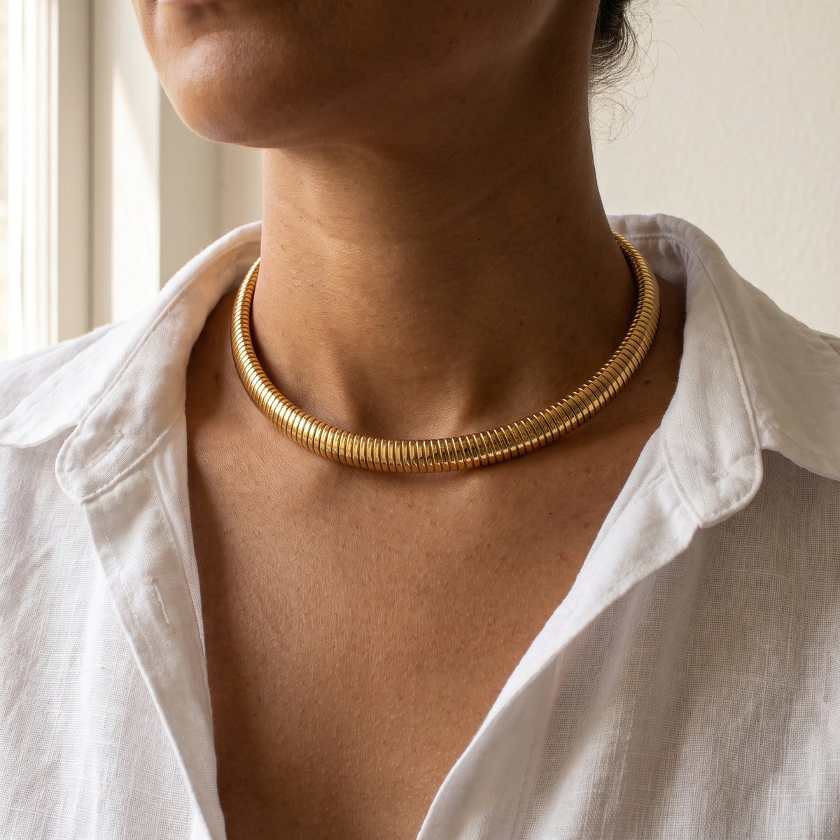 Collar Cala, gargantilla rígida de tubo dorado de acero bañado en oro, puesta con camisa blanca de lino