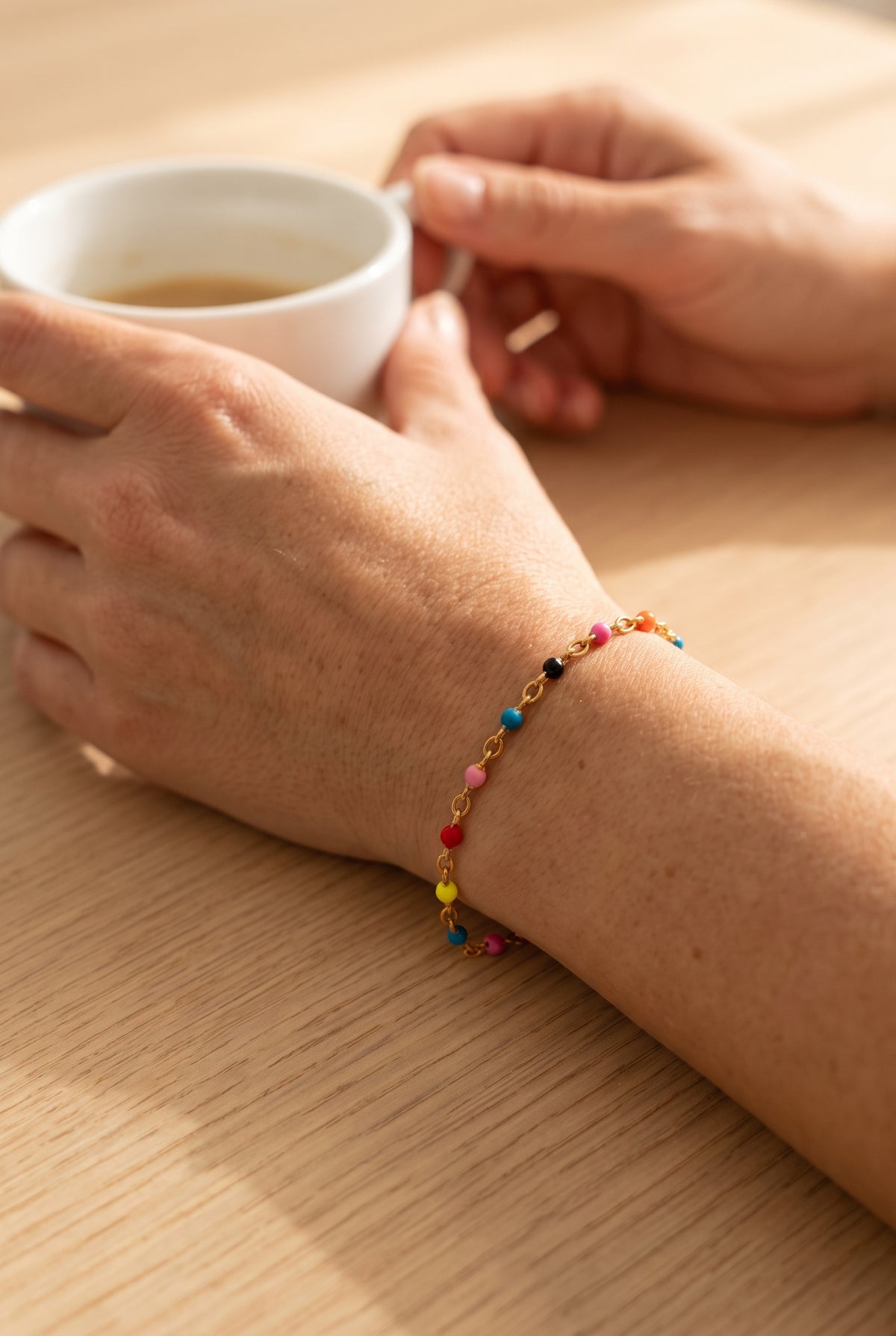 Muñeca con la Pulsera Maite, cadena dorada con cuentas de colores, sujetando una taza de café